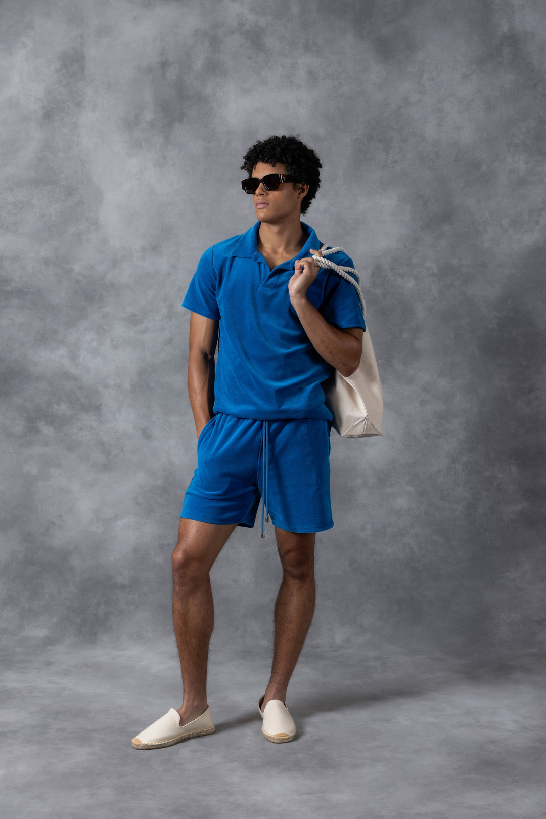 Model wearing Prince & Bond Riviera Terry cloth polo and shorts matching set in Mykonos Blue, styled with sunglasses on a neutral background